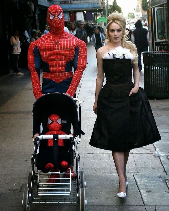 Spiderman in costume pushing a stroller with a child in matching outfit, walking beside a woman in a black dress on a city sidewalk.
