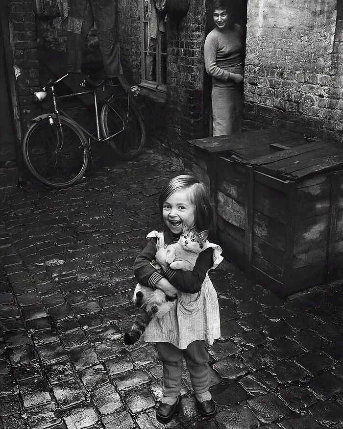 Little girl joyfully holding a cat on cobblestone street, with a woman smiling nearby in this fascinating vintage photo.
