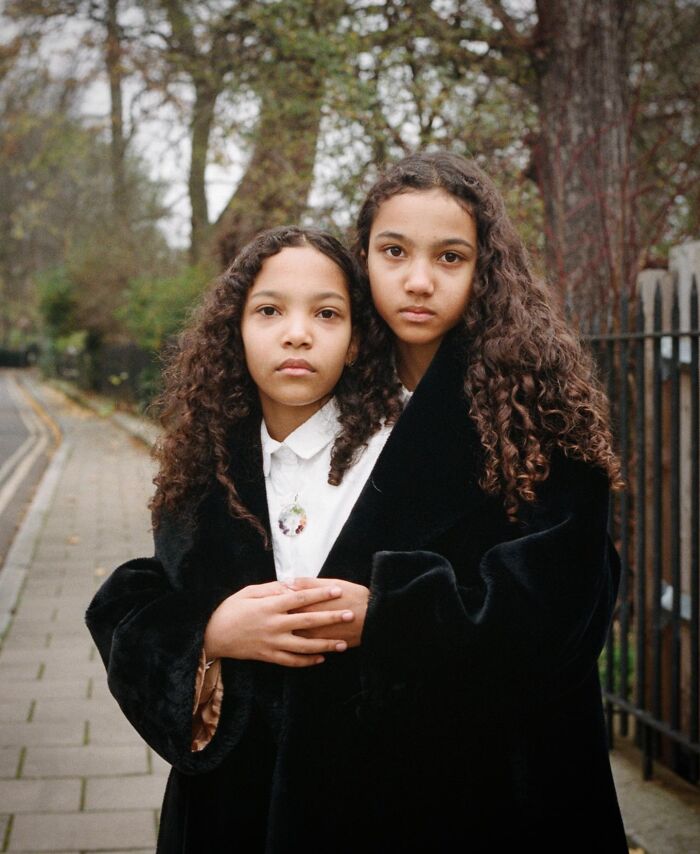 Twin girls with curly hair in black coats on a London street, captured by a London photographer in stunning portraits.