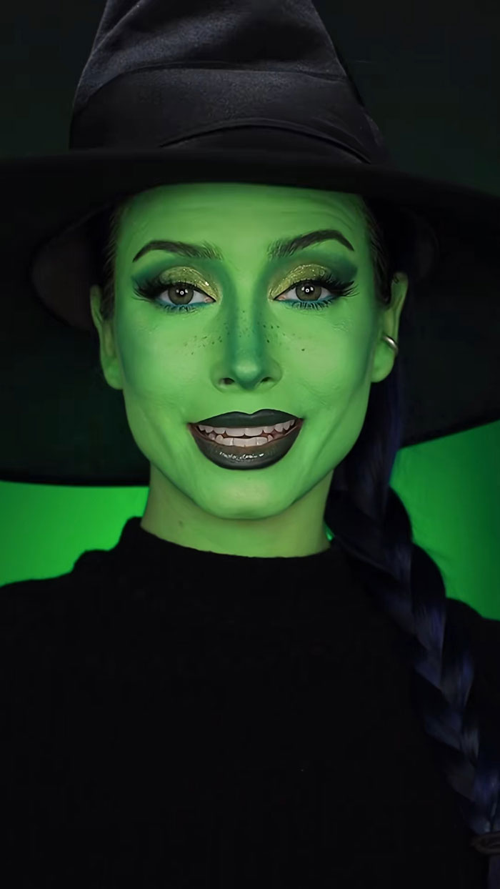 Woman with green Halloween makeup wearing a black witch hat and black outfit, showcasing creative Halloween makeup art.