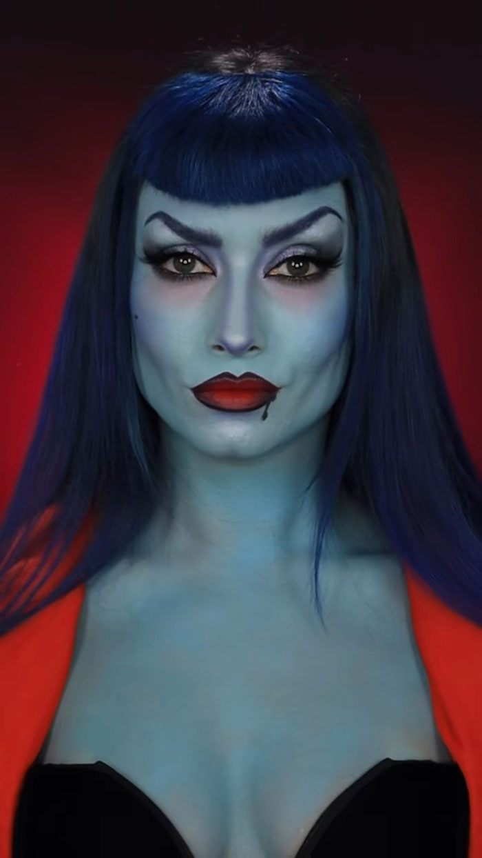 Woman with blue skin and dark blue hair wearing creative Halloween makeup and red lipstick against a dark red background.