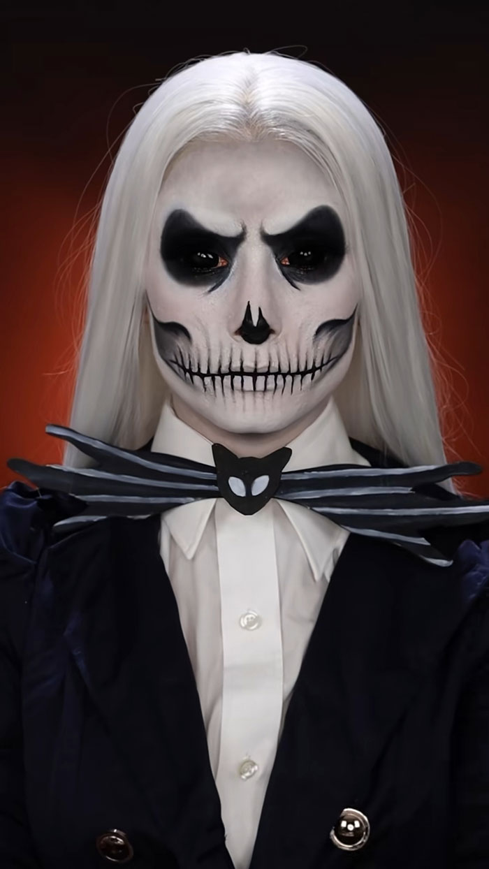 Person with Halloween makeup resembling a skeleton, wearing white shirt, black jacket, and spider-themed bow tie.