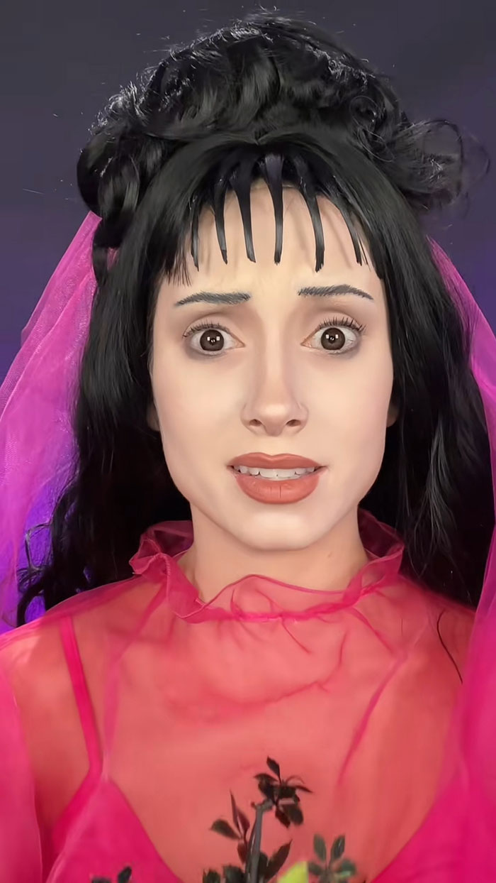Young woman with creative Halloween makeup wearing a pink sheer outfit and black styled hair against dark background