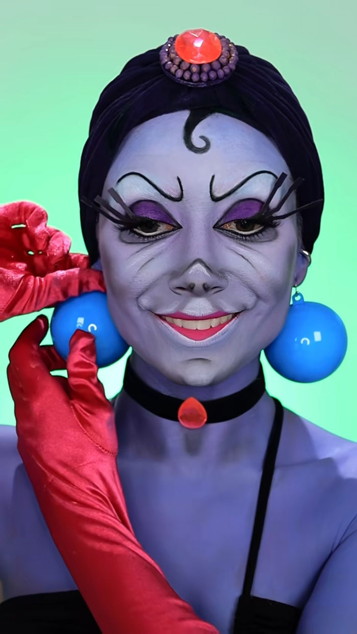 Creative Cliche Halloween makeup with purple face paint, dramatic lashes, red gloves, and blue ball earrings against a green background.