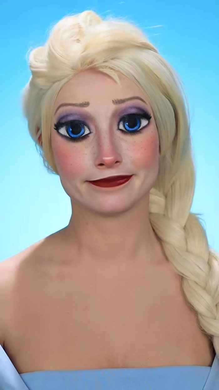 Creative Halloween makeup on woman with large blue eyes, blonde braided hair, and purple eyeshadow against blue background.