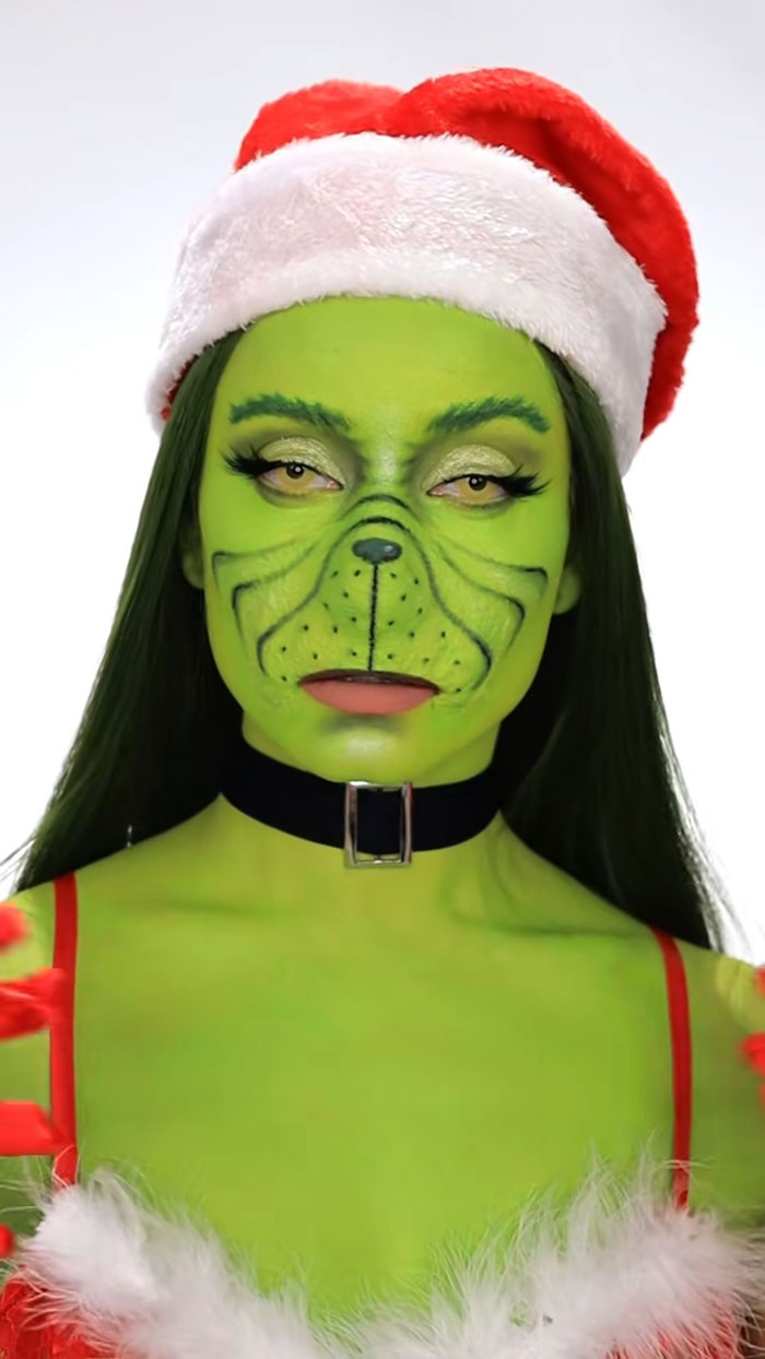 Woman with green Halloween makeup as a Grinch character wearing a Santa hat and festive outfit, showcasing creative Halloween makeup.