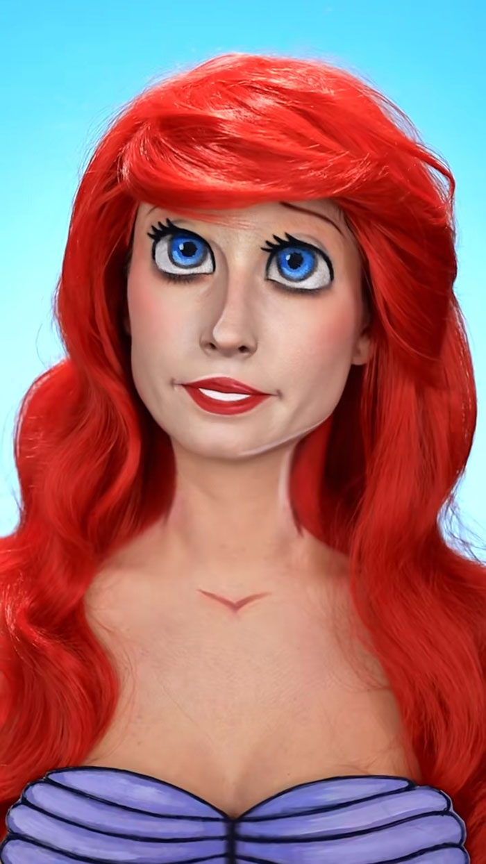 Woman with creative Halloween makeup and bright red wig, styled to resemble a cartoon character with large blue eyes.