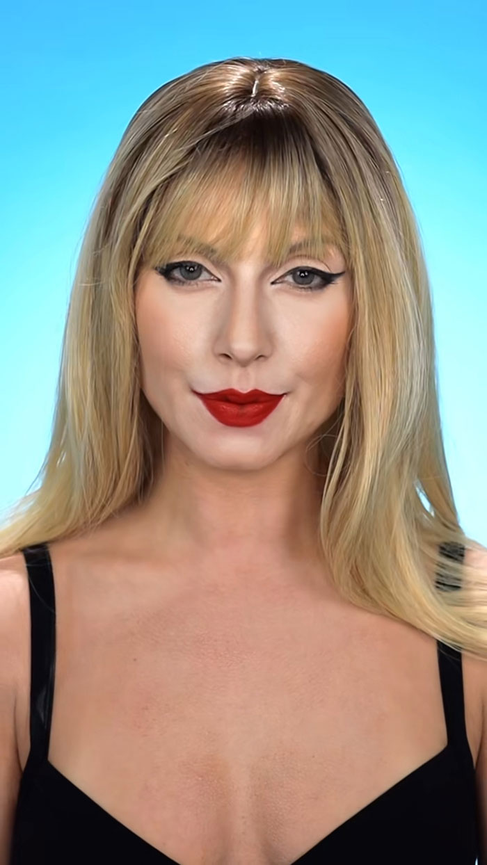 Blonde woman with striking Halloween makeup featuring winged eyeliner and bold red lipstick against a blue background