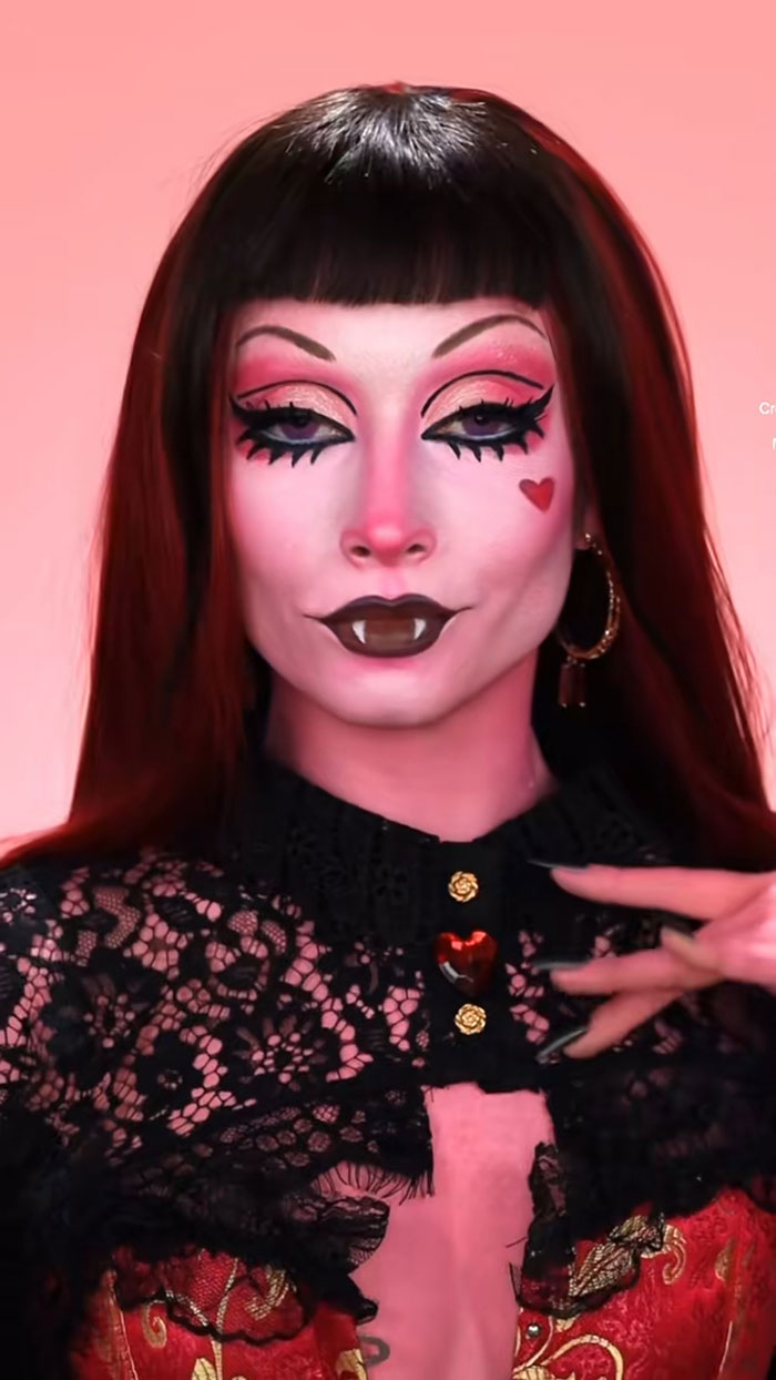 Creative Cliche with amazing Halloween makeup featuring vampire fangs, heart detail, bold eye makeup, and black lace costume.