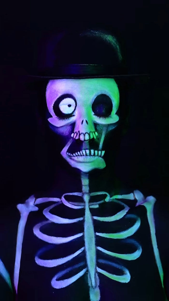 Creative cliche Halloween makeup of glowing skeleton with black hat on dark background showing artistic body paint.
