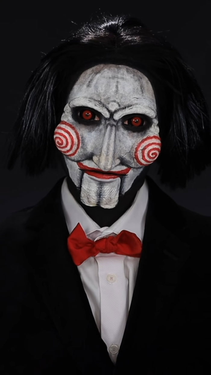 Person wearing amazing Halloween makeup as a creepy puppet with red eyes, white face paint, and a red bow tie in a dark suit.