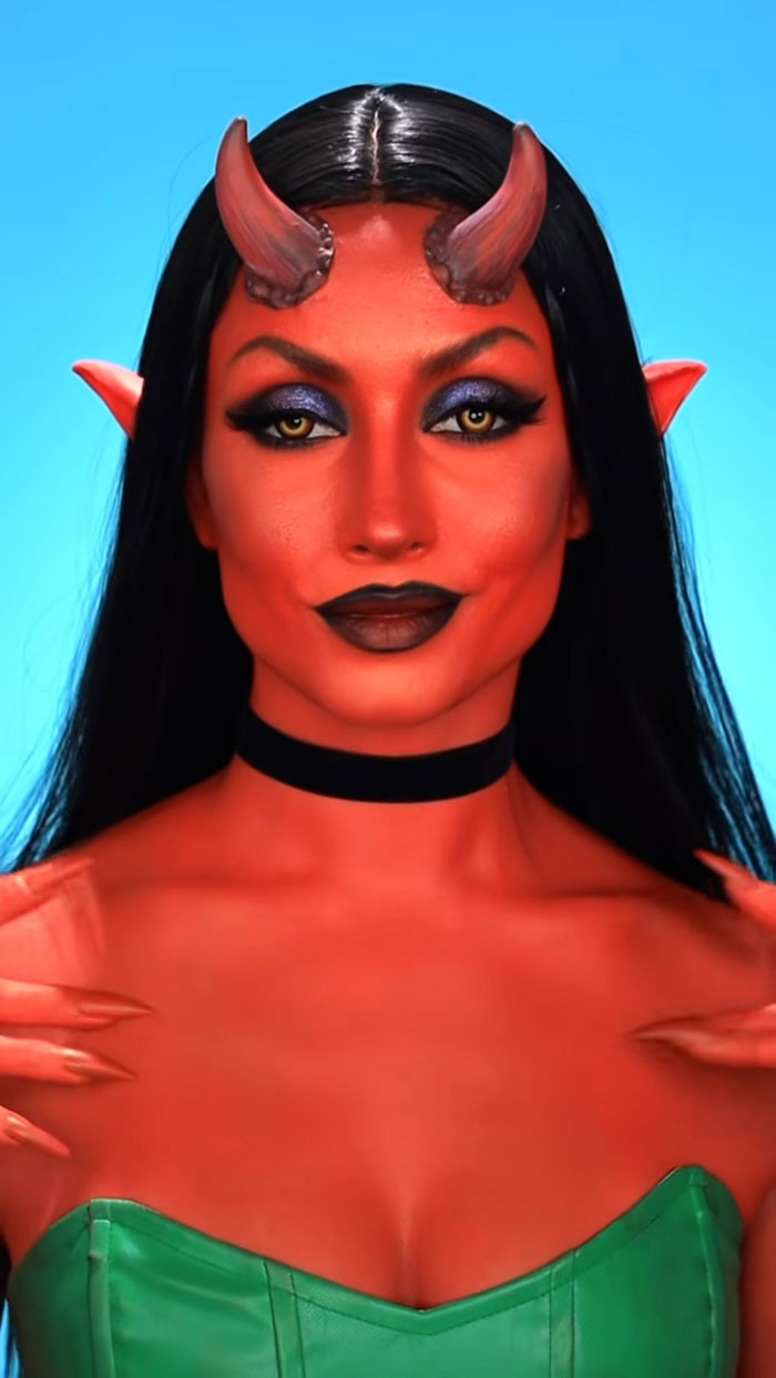 Woman with creative Halloween makeup as a red demon with horns, yellow eyes, and black hair wearing a green top against blue background