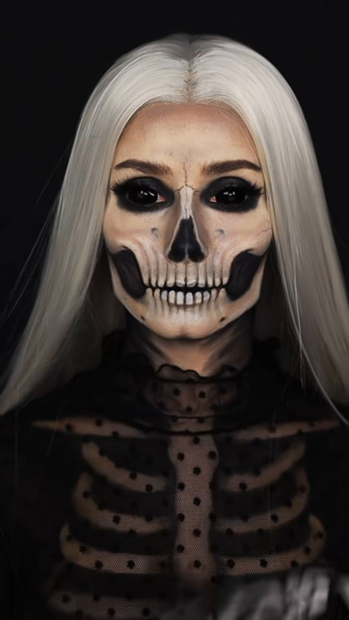 Woman with creative Halloween makeup showing detailed skeleton face and black skeleton outfit against dark background