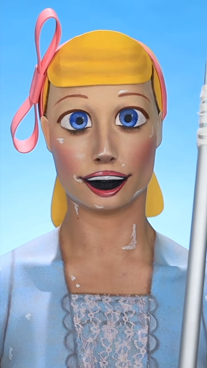 Halloween makeup transforming a person into a life-like doll with bright blue eyes, blonde hair, and a pink bow.