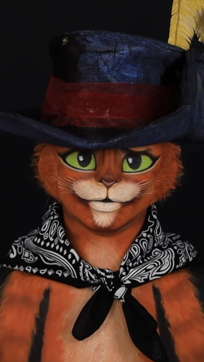 Creative cliche Halloween makeup transforming a person into a detailed orange cat wearing a hat and bandana.