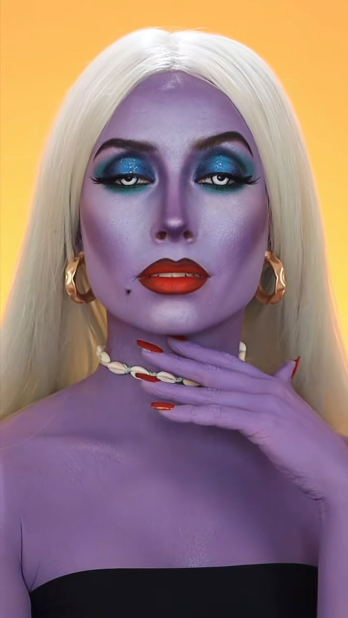 Creative Cliche Halloween makeup featuring purple skin, bold blue eyeshadow, red lips, and platinum blonde hair on a model.