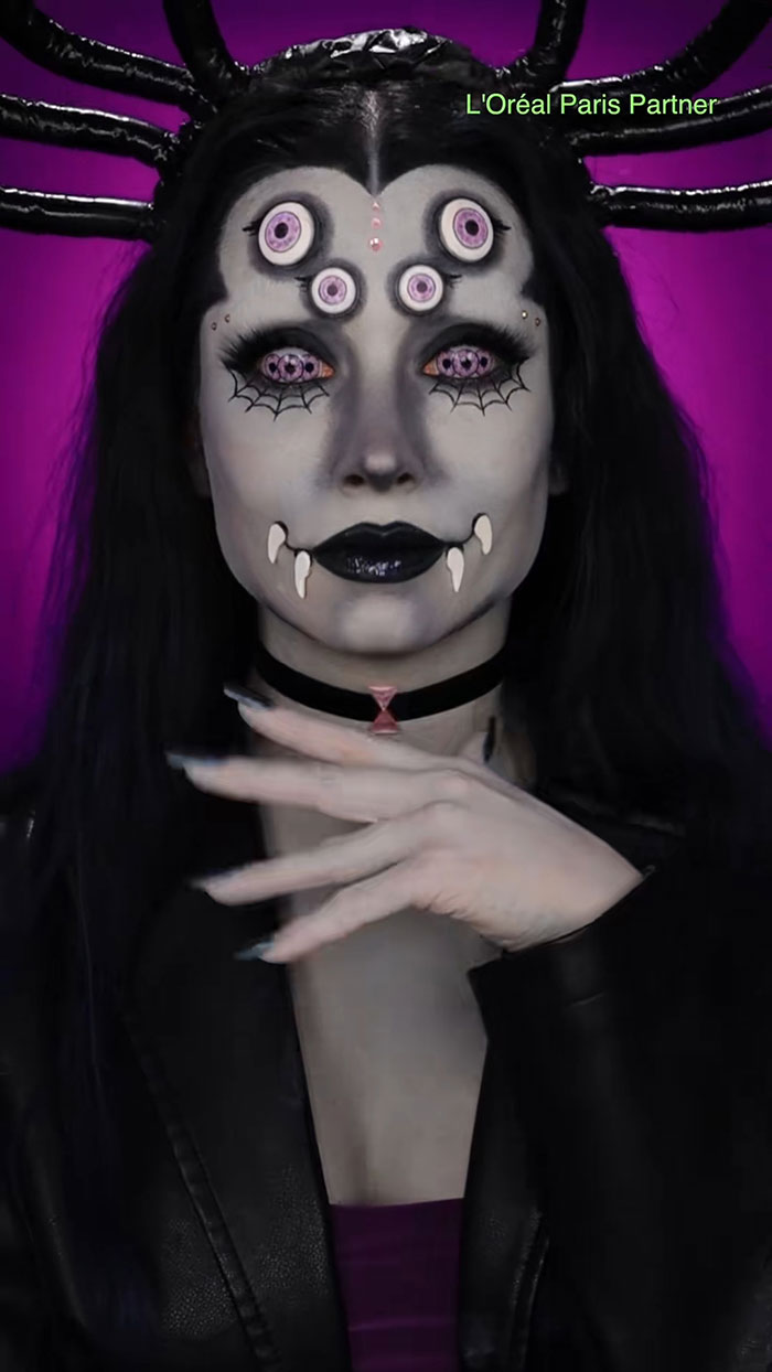 Halloween makeup showing a woman with spider-themed face paint, multiple eyes, fangs, and black spider legs on a purple background.
