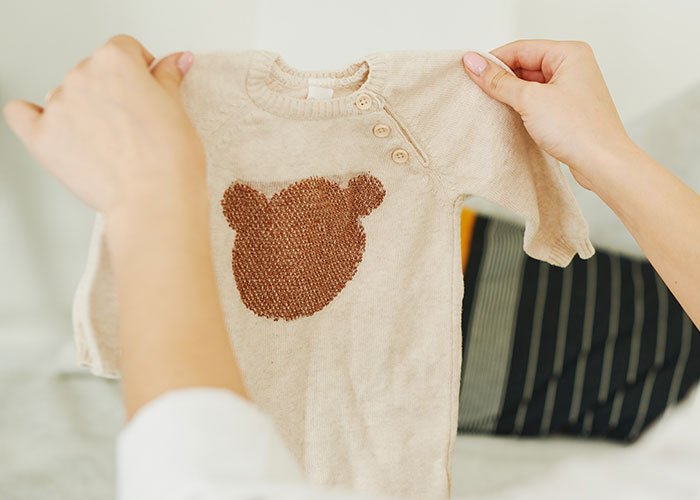 Hands holding a small beige baby outfit with a brown bear design, illustrating a wife leaving due to husband refusing to stand up.