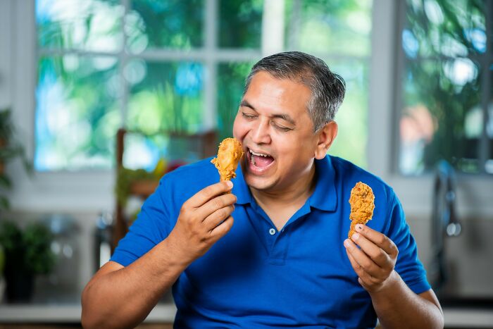 Man in blue shirt smiling and holding fried chicken drumsticks, highlighting healthy habits people are tired of.