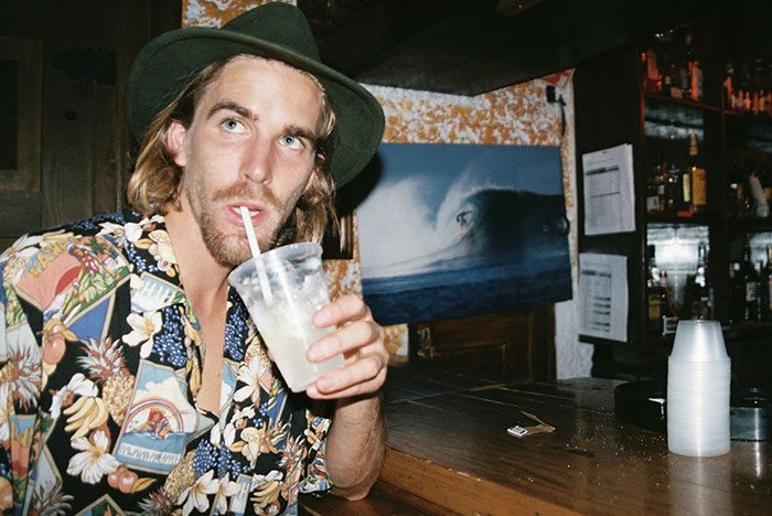 Man in a green hat and floral shirt drinking a beverage at a bar with instant karma theme in the background.