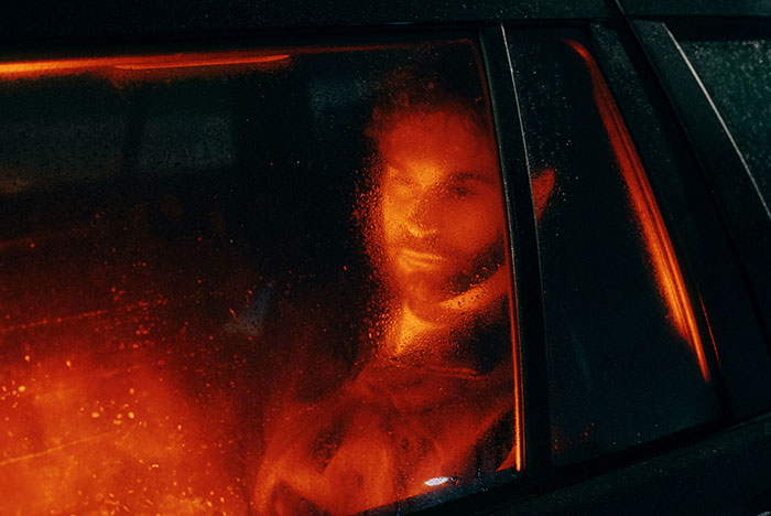 Man’s face illuminated by orange light, seen through a wet car window, illustrating instant karma moment.