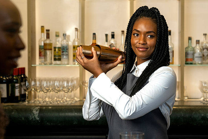 Young bartender shaking cocktail shaker behind bar, illustrating instant karma moment in a busy setting.