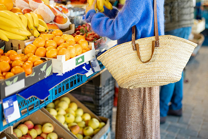 Person shopping at a fruit market with a large straw bag, surrounded by fresh bananas and other colorful produce.