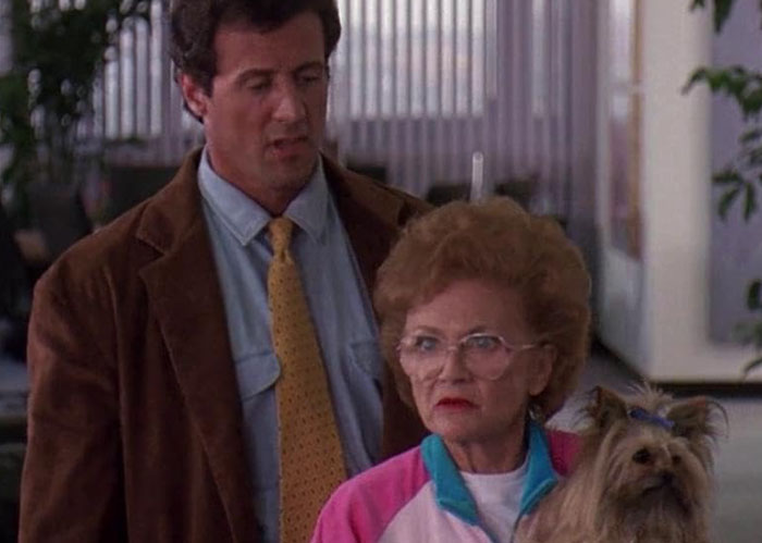 Man in brown jacket and yellow tie standing behind elderly woman with glasses holding a small dog in an office hallway.