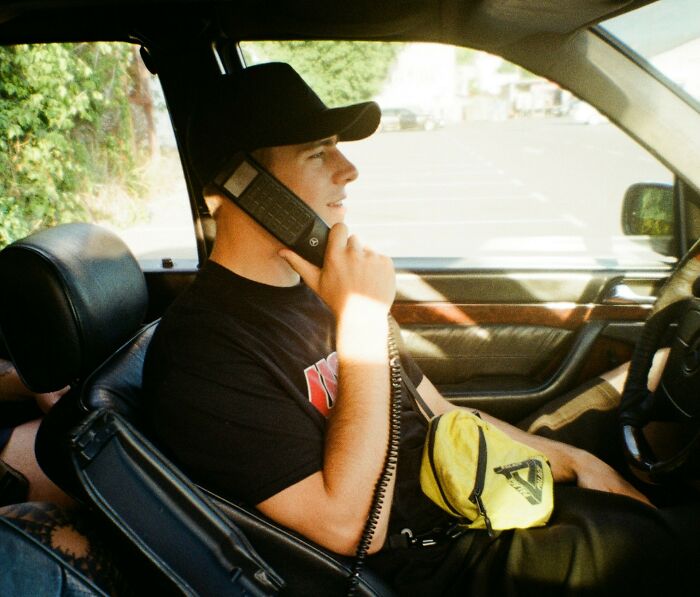 Young man in a car wearing a cap and holding a retro phone, capturing a chaotic breakup story moment.