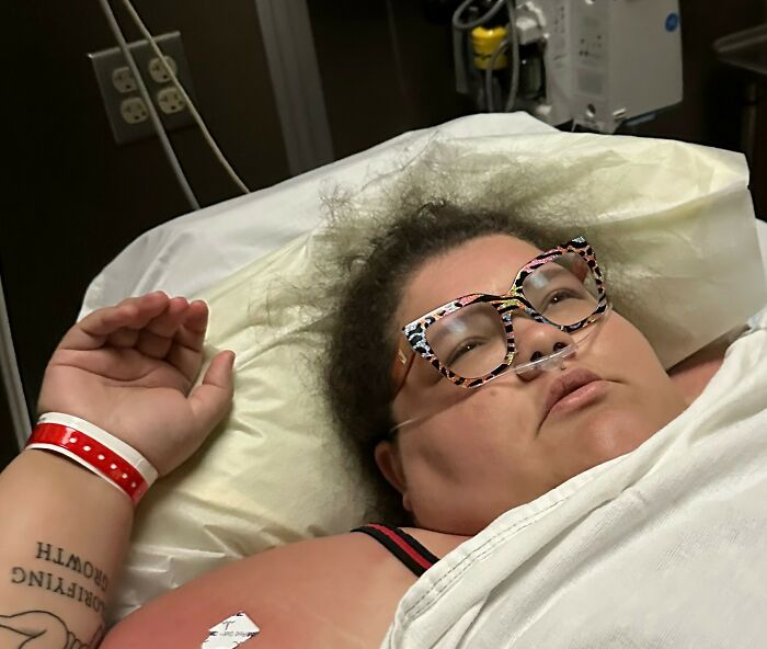 Plus-size activist lying in a hospital bed with oxygen tube and patterned glasses, appearing to rest after a health issue. Plus-size activist lying in a hospital bed with oxygen tube and patterned glasses, appearing to rest after a health issue.