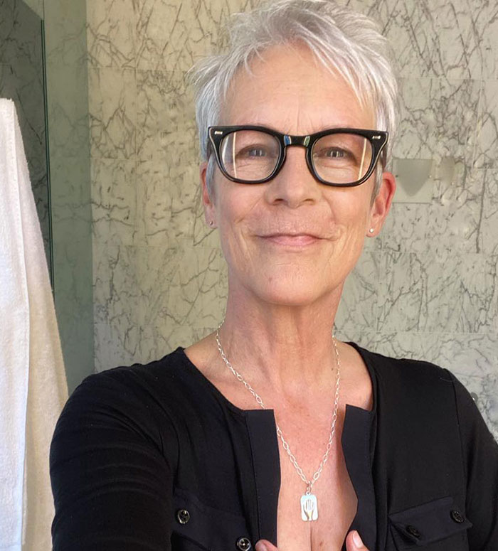 Jamie Lee Curtis smiling, wearing glasses and a black top, reacting to Sydney Sweeney&rsquo;s braless look stirring online drama.