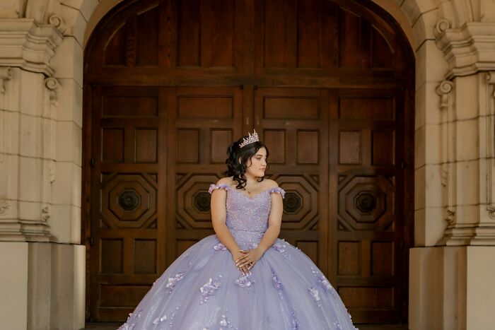 Teen girl in a purple gown and tiara standing alone by wooden doors, reflecting on popular kids in school outcasts.