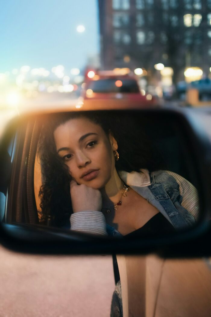 Young woman with curly hair looking thoughtful in a car side mirror at dusk, reflecting on drunk fumbles and bad decisions.