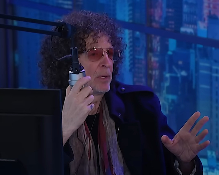 Man with curly hair and sunglasses speaking into a microphone during an online podcast about Jennifer Lopez cheating accusations.