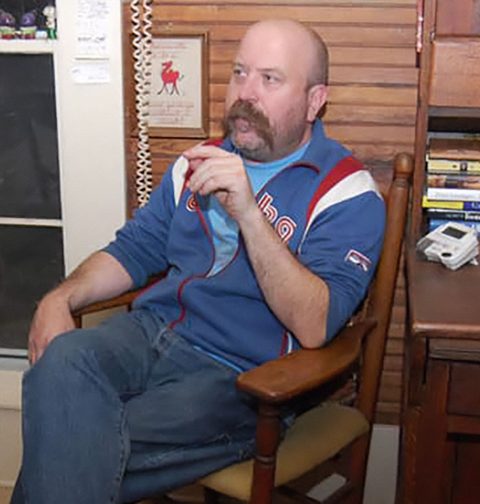 Hombre sentado en silla de madera, con chaqueta azul, en un interior, relacionado con figuras públicas desaparecidas sin rastro.