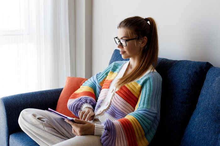 Young woman wearing glasses and a colorful sweater sitting on a couch using a tablet, illustrating anxiety issues.