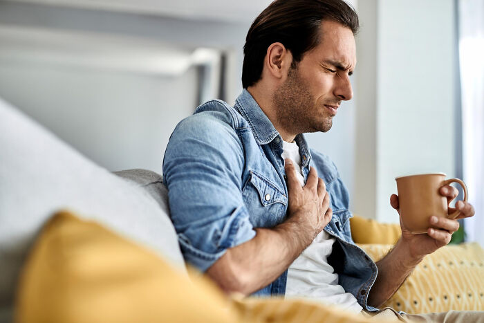 Man in denim jacket holding chest in pain while sitting on couch with a cup, illustrating anxiety misdiagnosis issues.