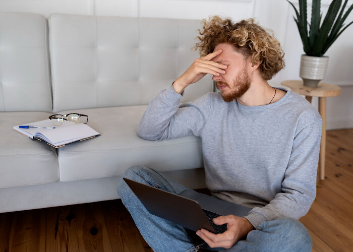 Man sitting on floor holding laptop, appearing stressed and anxious while working in a casual home setting.