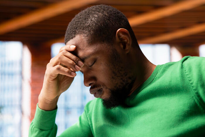 Man in green sweater holding forehead, appearing stressed and anxious while sitting indoors near windows.