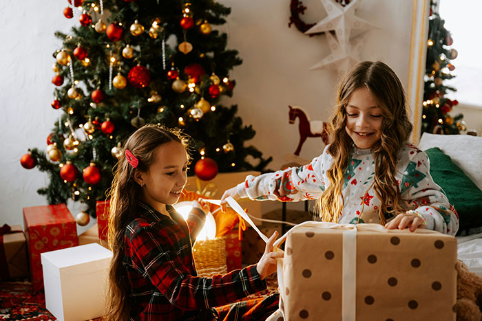 Two girls happily opening presents by a decorated Christmas tree, capturing a family moment with kid&rsquo;s photos and gifts.