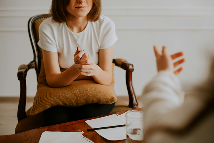 Woman with folded hands sitting on a chair talking to another person during a counseling session indoors
