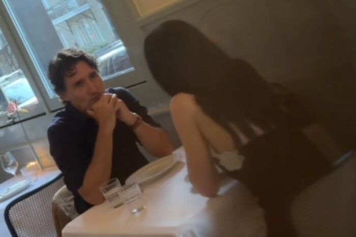 Katy Perry and shirtless Justin Trudeau sitting and talking closely at a cozy indoor table setting.