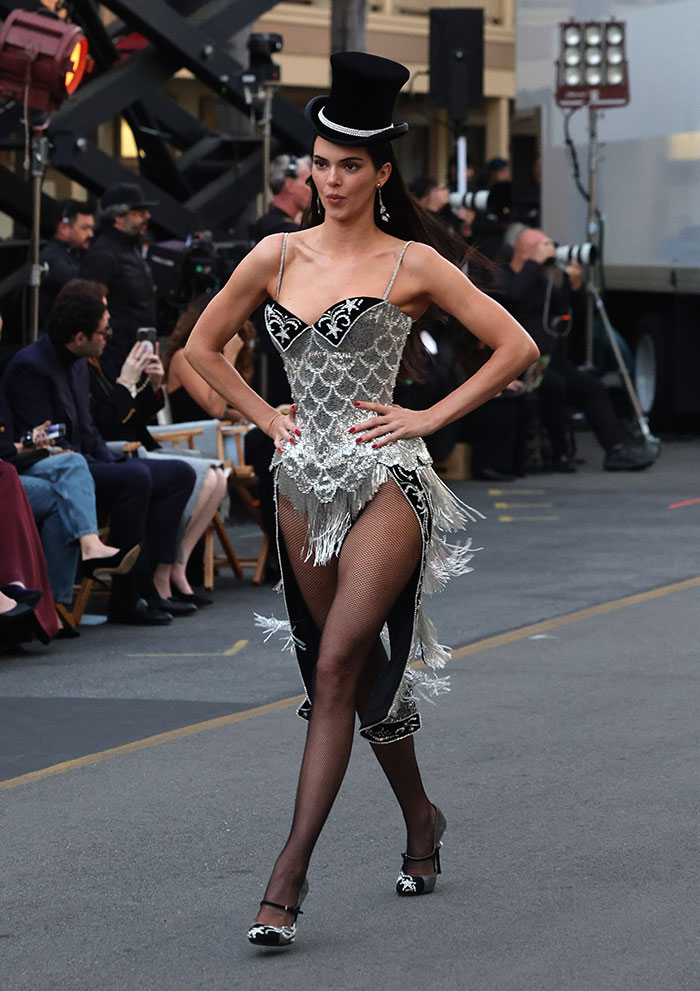 Model in a silver and black intricate dress with fishnet stockings and a top hat at Vogue World Hollywood 2025 celebrity looks event.