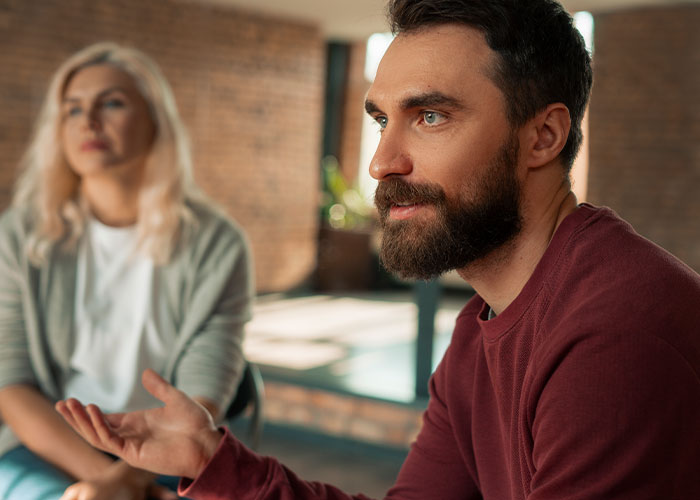 Man with beard speaking seriously while woman in background looks upset during family discussion about conservative uncle.
