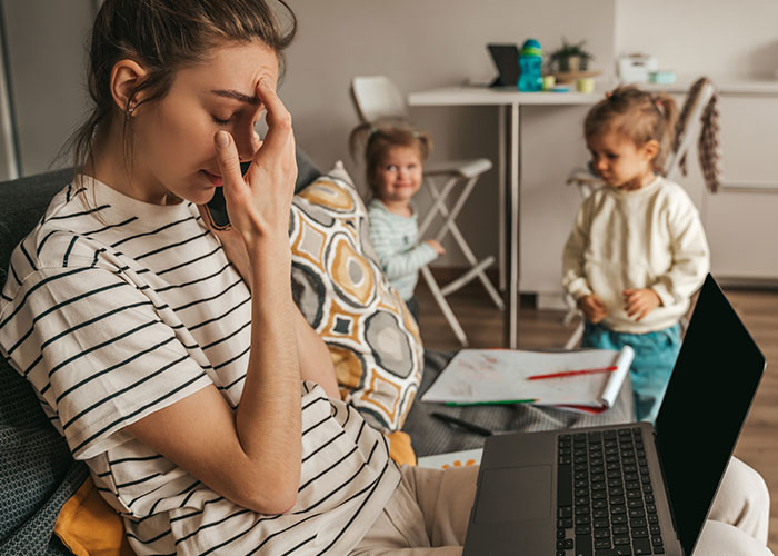 Stressed woman working on a laptop at home while managing kids, illustrating challenges of kicking out sister&rsquo;s kids during a family emergency.