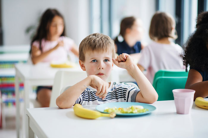 Young boy left hungry at school, sitting with untouched snack and thoughtful expression during lunchtime.