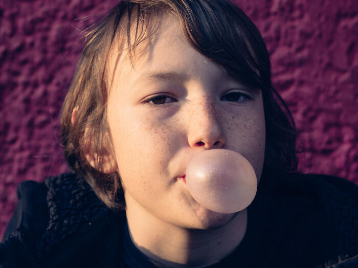 Child with freckles blowing a bubble with gum, illustrating random childhood things people now realize weren’t normal.