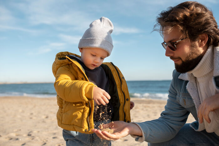 Toddler in yellow jacket playing with sand on the beach with an adult, illustrating random things people did as kids.