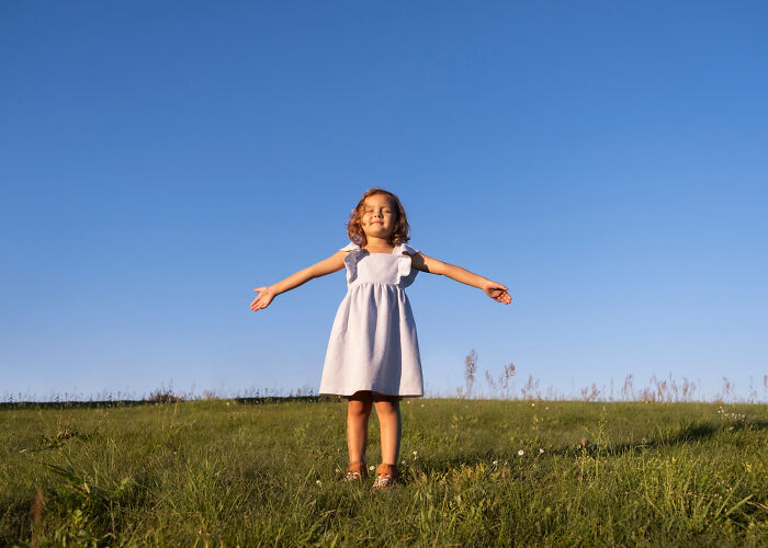 Young girl standing with arms outstretched on grass under clear blue sky, representing quirky things kids did.