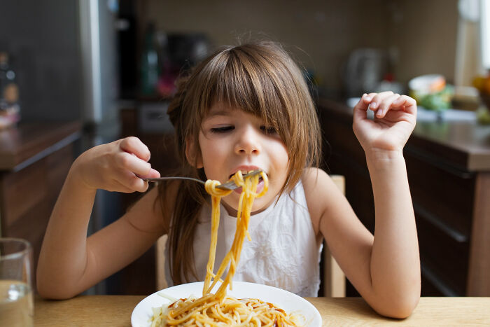 Young girl eating spaghetti at home, showcasing a random childhood moment people only now realize wasn’t normal.